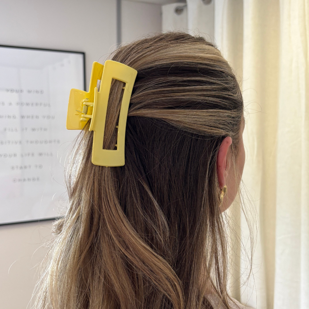 Blonde woman wears a yellow square claw clip, holding her hair back, standing indoors near a window.