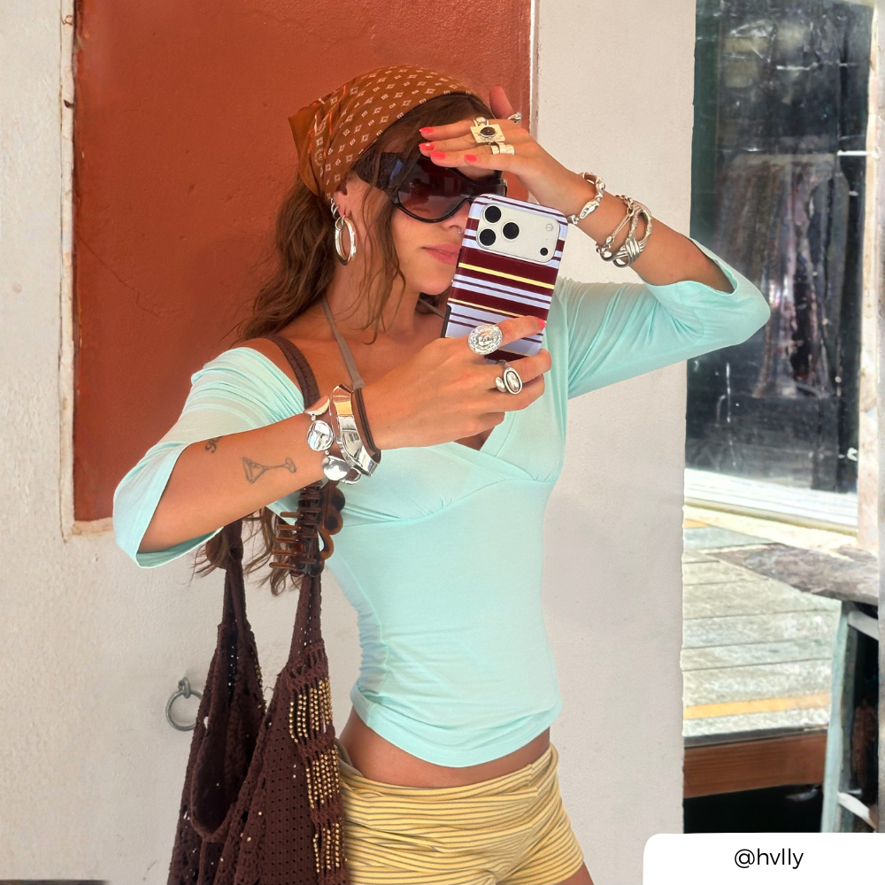 A woman takes a mirror selfie wearing a light mint green off-the-shoulder crop top, yellow striped shorts, a brown patterned headscarf, and multiple gold rings and bracelets. She is holding a brown, blue and yellow striped phone case and carrying a brown woven bag against an outdoor storefront background.