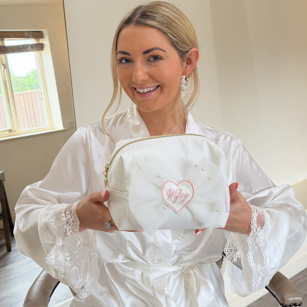 Bride in a white robe holding a white makeup bag with 'Wifey' printed on it.