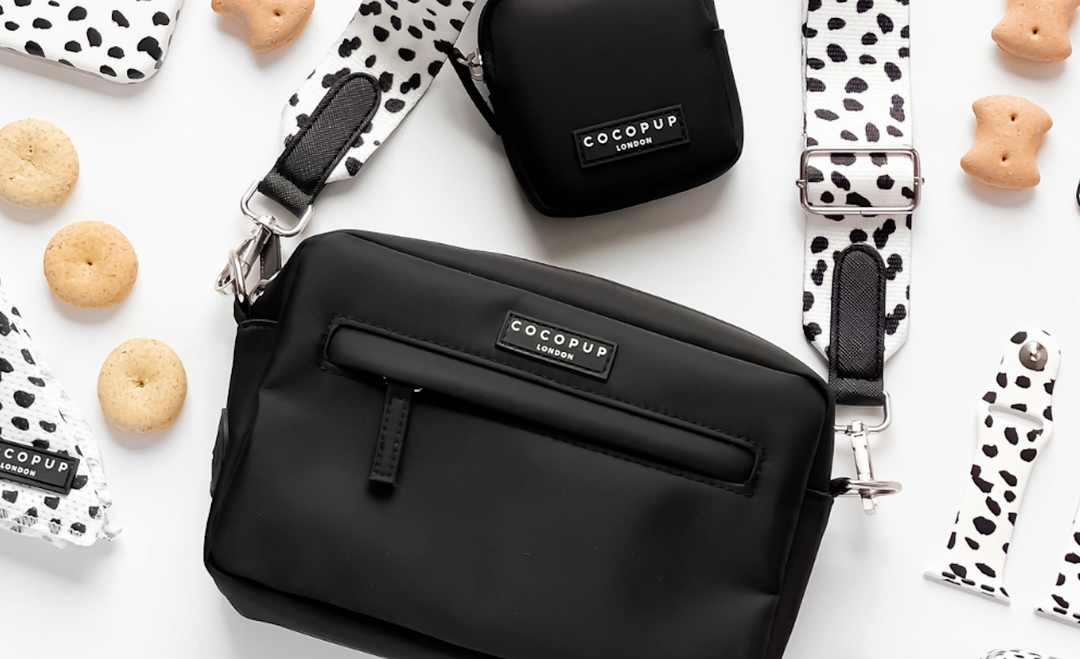 A stylish collection of black bags with matching Dalmatian-print straps is displayed on a white background. Surrounding the bags are Dalmatian-print accessories and dog treats.