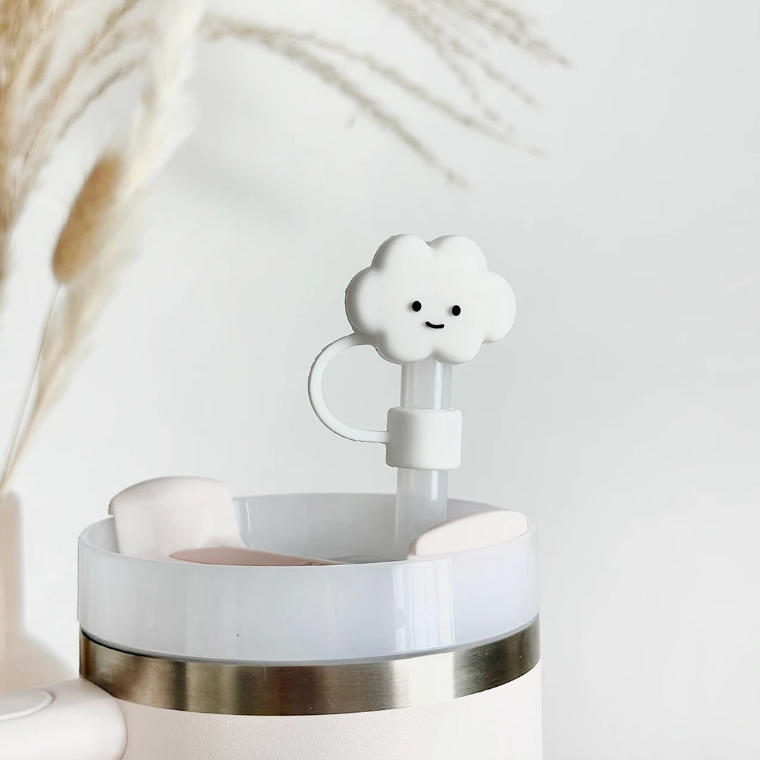 A white reusable cup with a light blue lid featuring a cute cloud-shaped stopper attached to a straw. The background includes blurred, decorative dried plants.