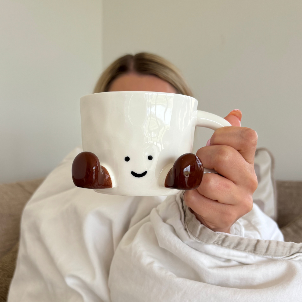 A person wrapped in a white blanket holds up a white mug with a cute smiling face and brown feet, partially obscuring their face.