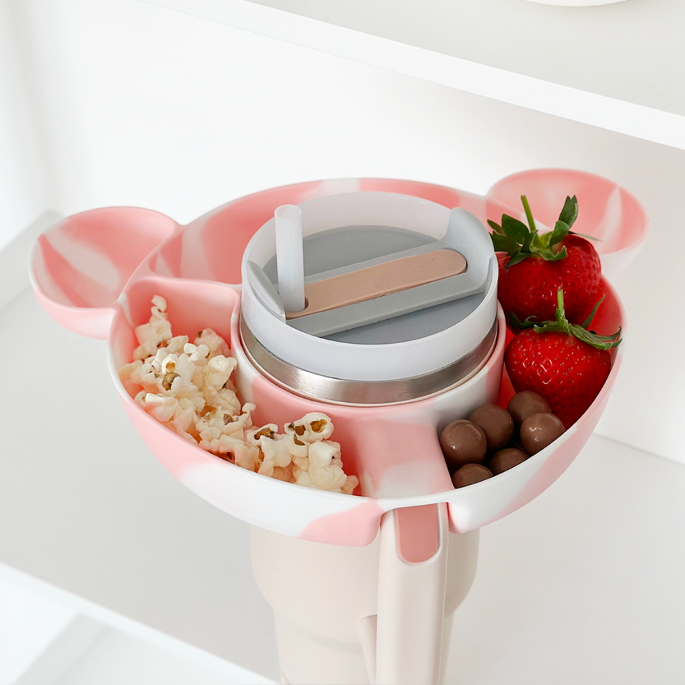 A pink and white Mickey Mouse-shaped tray with compartments holds popcorn, strawberries, chocolate-covered candies, and a lidded cup with a straw. The tray is placed on a white surface.