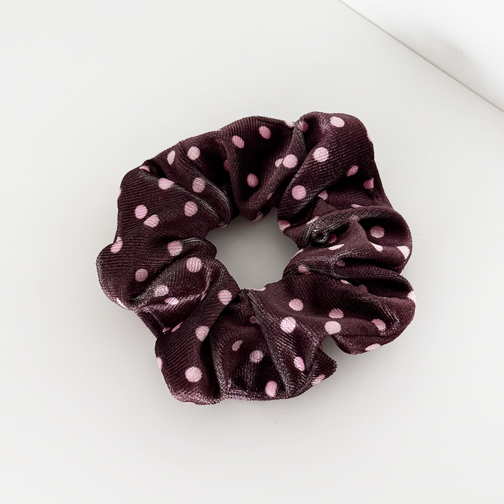 A dark brown velvet scrunchie with light pink polka dots, placed on a white surface near the corner of a wall.