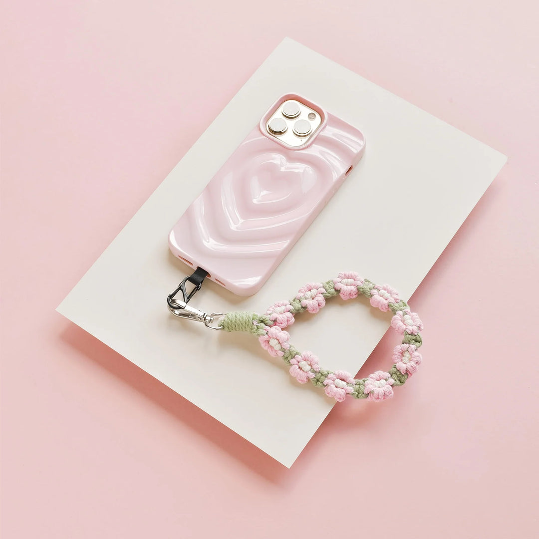 A pink smartphone case with a heart design lies on a white paper atop a pink surface. Attached is a crocheted pink and green flower keychain.