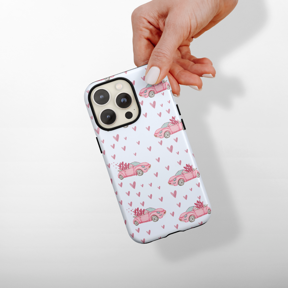 A hand with manicured nails holds an iPhone with a white case decorated with pink hearts and illustrations of pink cars carrying flowers.