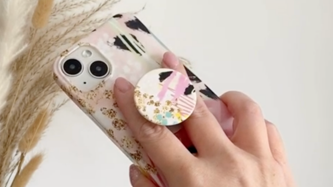 A hand holding a smartphone with a pink, abstract-patterned case and a pop socket adorned with colorful designs. In the background, dried decorative plants are visible against a neutral wall.