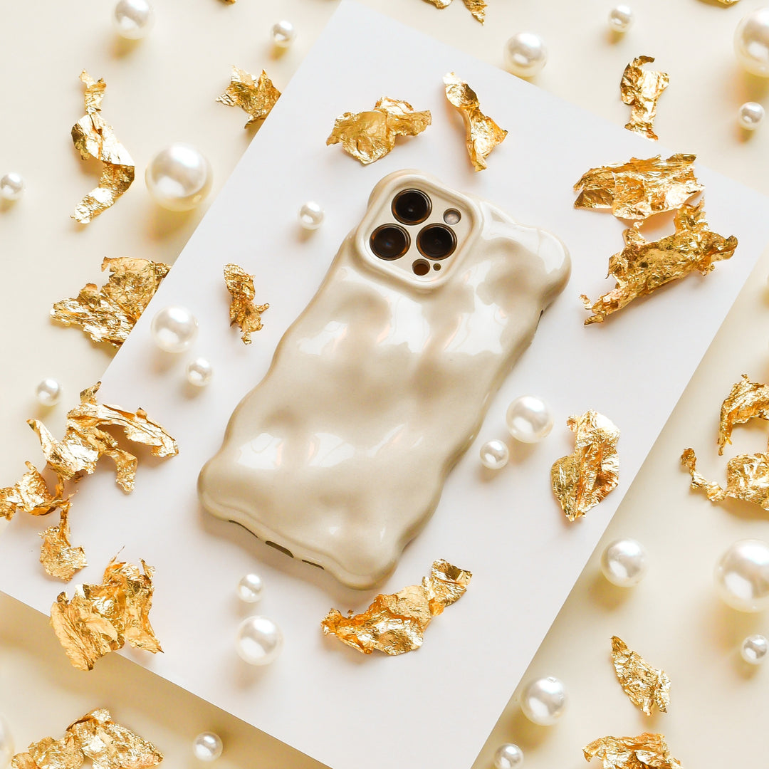 A wavy-textured gold phone case on a smartphone, surrounded by scattered gold foil and pearls on a cream background. The phone is placed on a white rectangular surface.