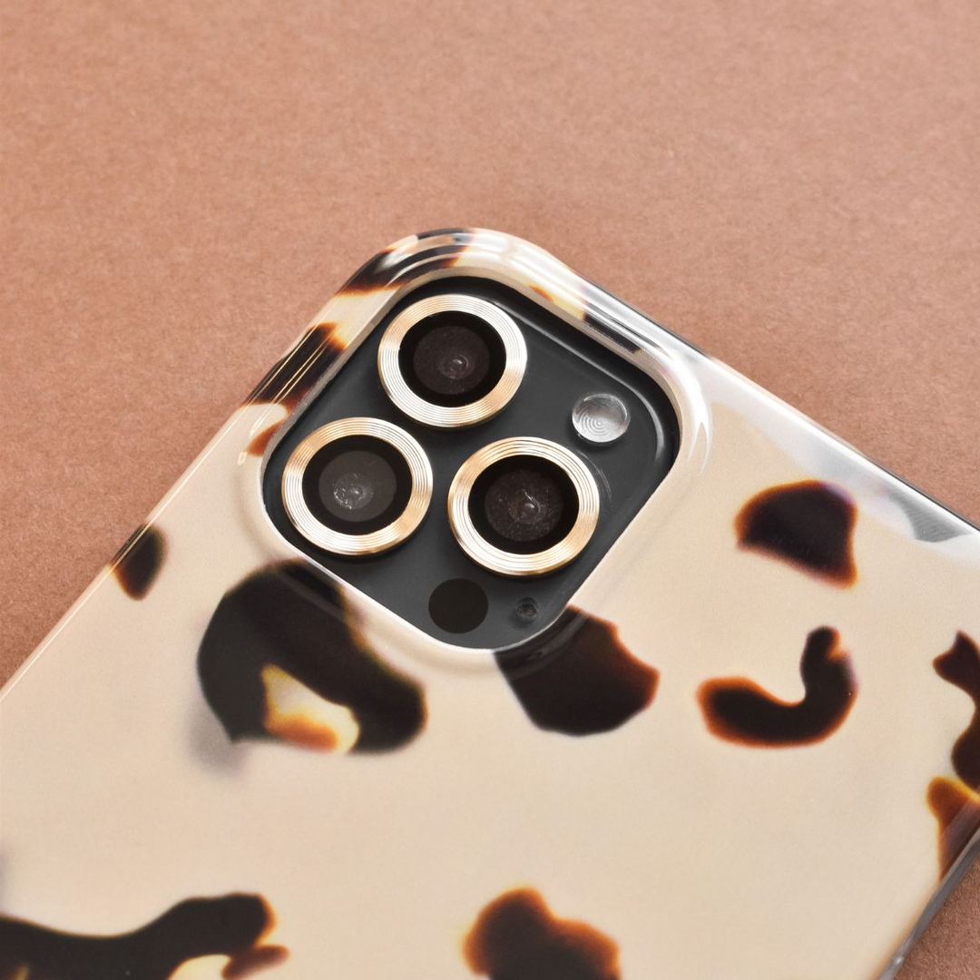 Close-up of a smartphone with a beige and black tortoiseshell patterned case, focusing on the triple camera lenses. The phone is placed on a brown surface.