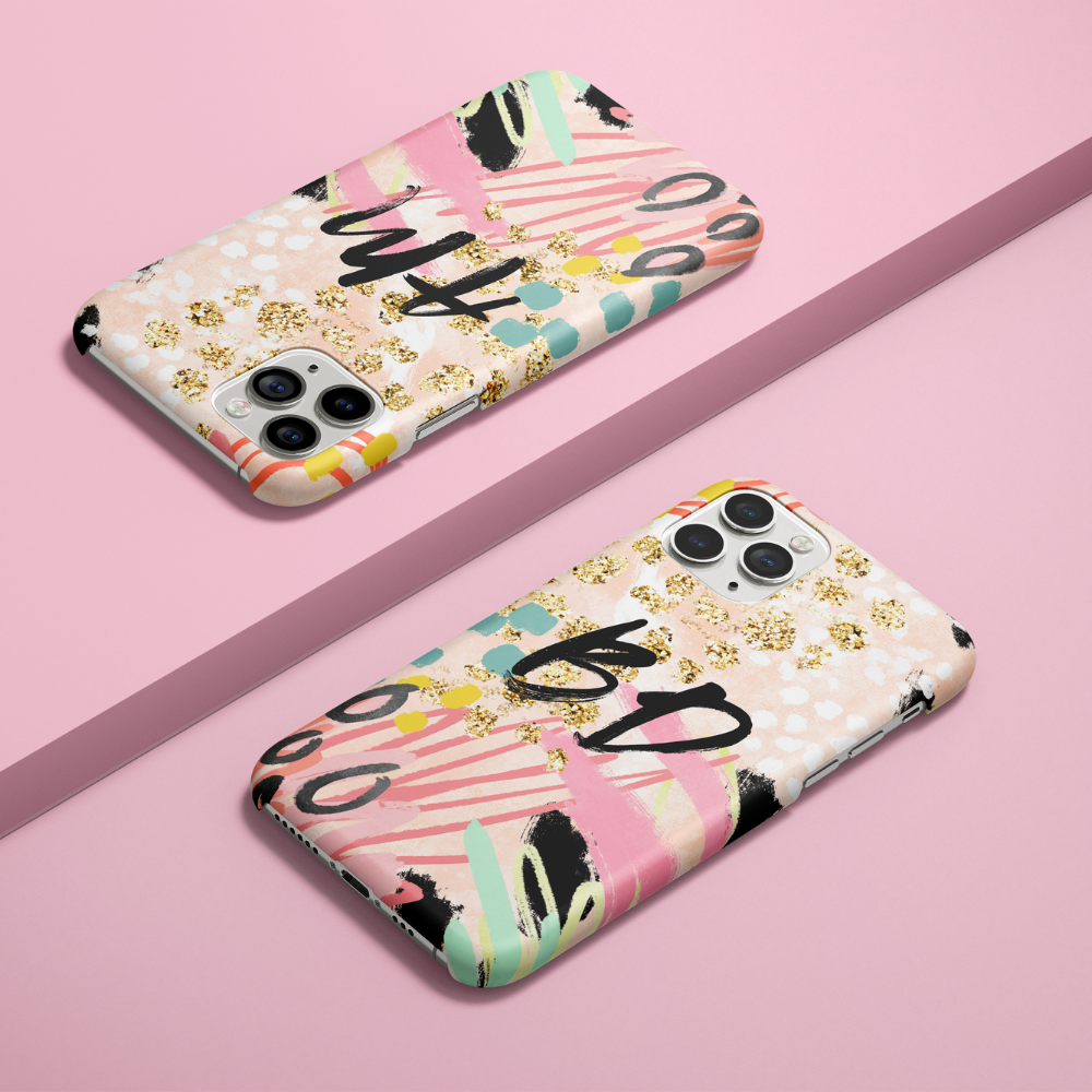 Two colorful, abstract-patterned smartphone cases with gold accents are displayed diagonally on a pink surface. Each case features a unique design of bold letters and brushstrokes.