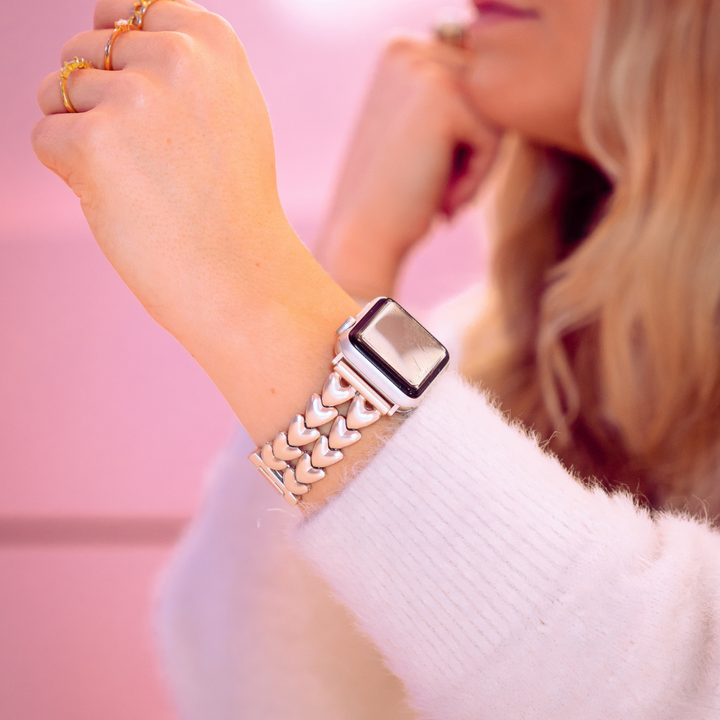 A person with long blonde hair, clad in a white fluffy sweater, is showcasing the Coconut Lane Hearts Chain Apple Watch Strap - Starlight on their wrist. The strap boasts a woven metal band, set against a soft pink gradient backdrop. Their hand is slightly raised, adorned with multiple rings.