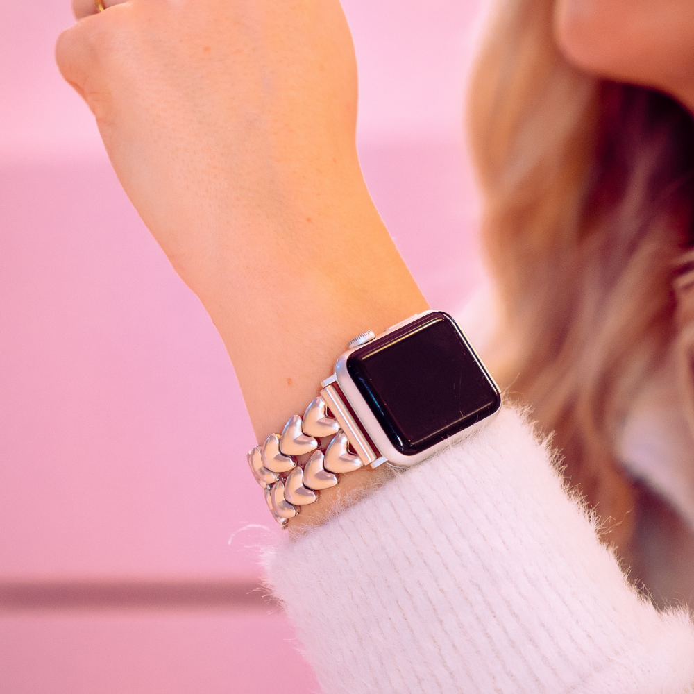 A close-up of a person's wrist wearing an Apple Watch with the Hearts Chain Apple Watch Strap - Starlight by Coconut Lane, featuring heart-shaped links. The person has a light skin tone, is dressed in a white fuzzy sweater, and the background is pink. This stainless steel adjustable strap fits ALL Apple Watch models.