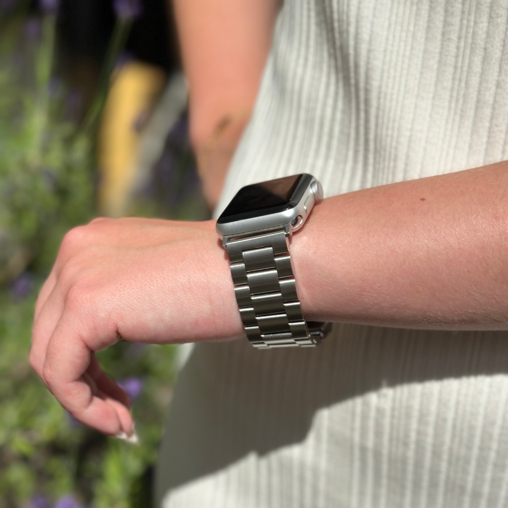 A person wearing a Coconut Lane Stainless Steel Apple Watch Strap in Silver on their left wrist. The person is dressed in a white ribbed sleeveless top and is standing outdoors with greenery and purple flowers blurred in the background.