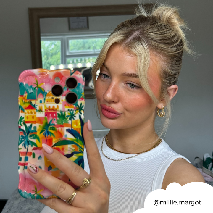 A young woman with blonde hair tied in a high bun takes a mirror selfie holding a phone adorned with the Portofino Dreams Wavy Phone Case by Coconut Lane. She is wearing a white sleeveless top, hoop earrings, and a gold necklace. Her Instagram handle "@millie.margot" is included in the bottom right corner.