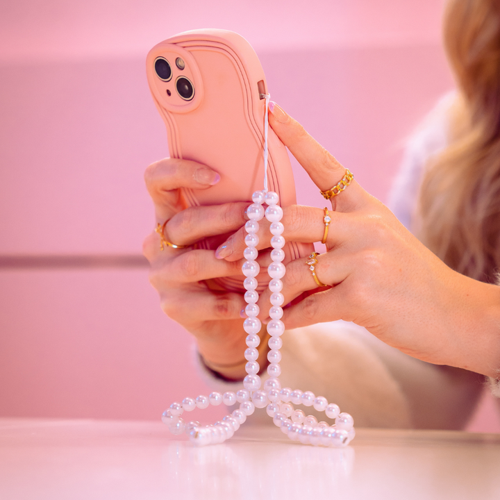 A person with blonde hair holds a pink smartphone adorned with a pearl beaded strap. The person is wearing several gold rings on their fingers, and the background is a soft pink color, showcasing the NAKD Curvy Phone Case - Strawberry by Coconut Lane that offers both style and protection.