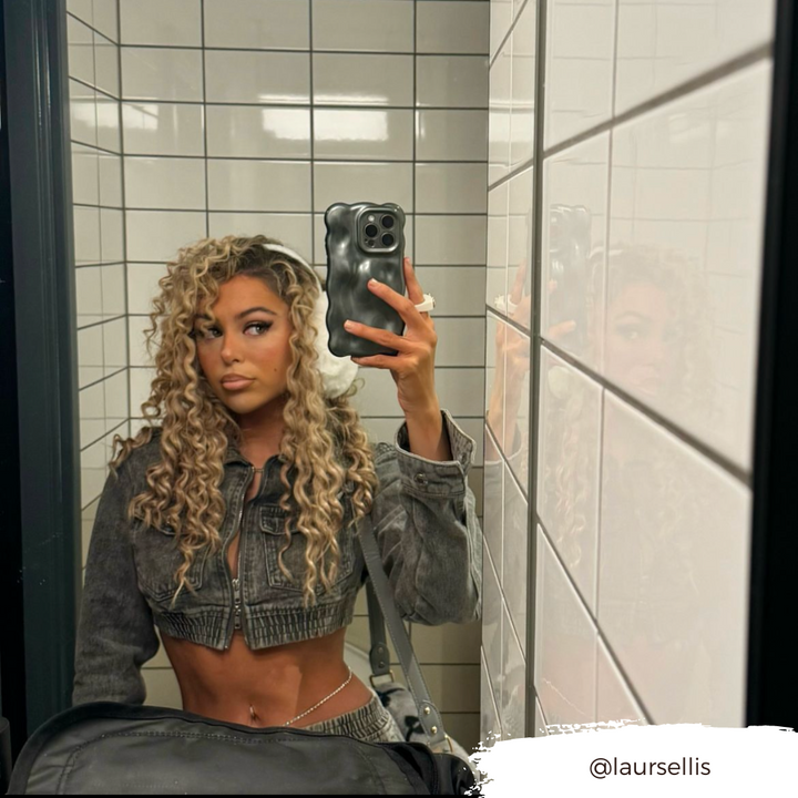 A woman with long, curly blonde hair takes a selfie in a tiled bathroom mirror. She's wearing a cropped denim jacket exposing her midriff and holds her phone, which is encased in an extra protective Coconut Lane 3D Pearl Phone Case in grey. Her expression is neutral. The Instagram handle @laursellis is visible in the bottom right corner.