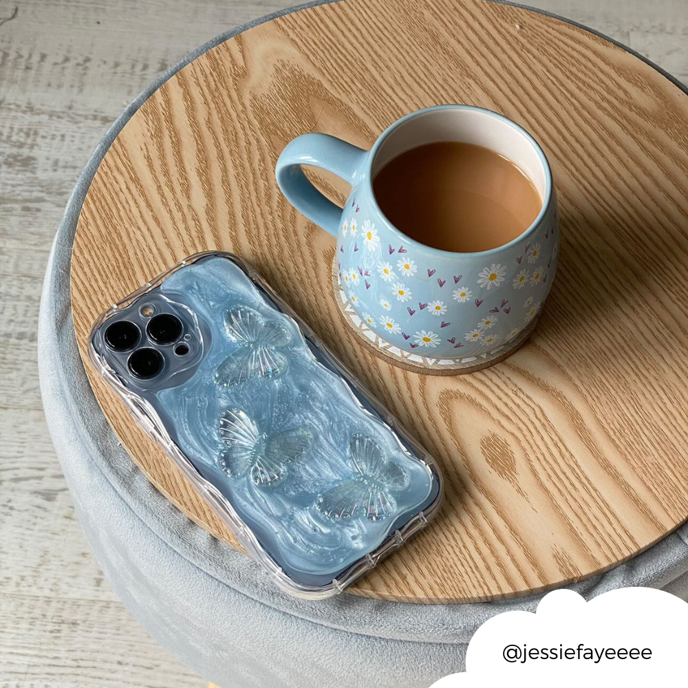 A blue mug with floral designs filled with tea sits on a round wooden table. Beside it is an iPhone encased in the Shimmer Blue Butterflies Phone Case by Coconut Lane. A white circular tag in the bottom right corner shows the handle @jessiefayeeee.