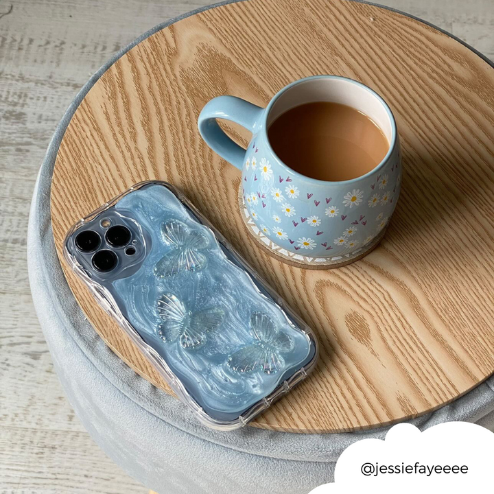 A blue mug with floral designs filled with tea sits on a round wooden table. Beside it is an iPhone encased in the Shimmer Blue Butterflies Phone Case by Coconut Lane. A white circular tag in the bottom right corner shows the handle @jessiefayeeee.