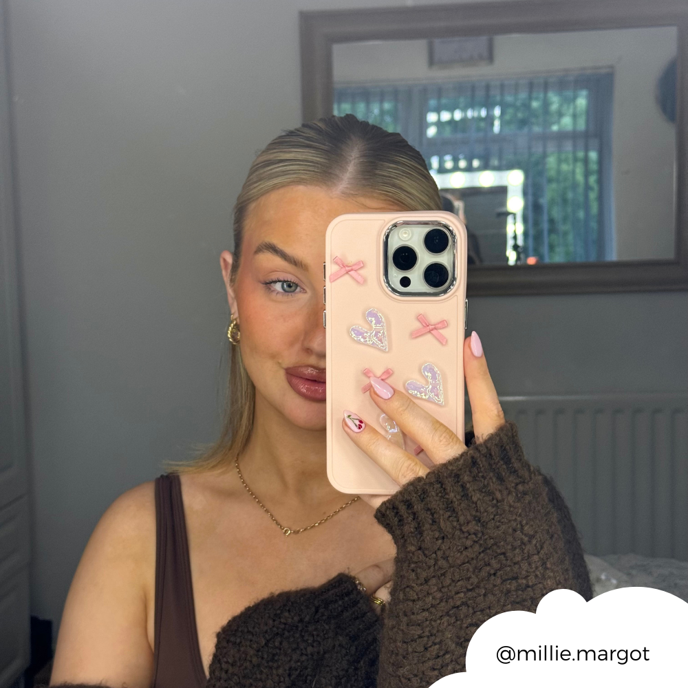 A woman with blonde hair, dressed in a brown outfit, takes a mirror selfie with her iPhone15 encased in the Coconut Lane Pearly Hearts Phone Case - Pink, featuring heart and x designs. She stands in a room where the white wall and window reflect greenery. The photo is shared via the handle @millie.margot.