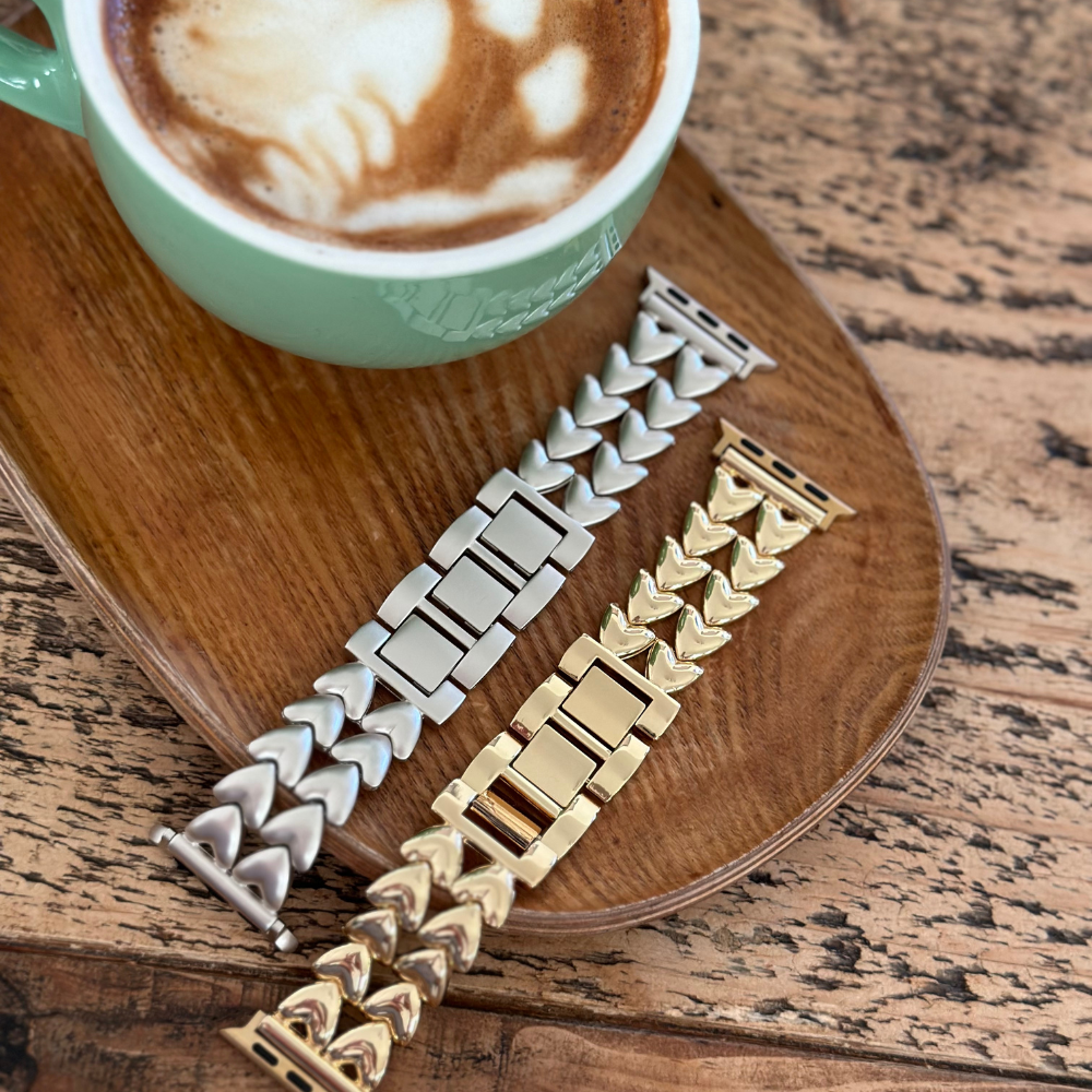 A green cup of cappuccino sits on a wooden tray on a rustic wooden table. Next to it are two watch bands: one silver with a unique chunky link design and one gold with a similar style. These Coconut Lane Hearts Chain Apple Watch Straps in Starlight fit ALL Apple Watch models, adding to the cozy, stylish vibe of the setting.