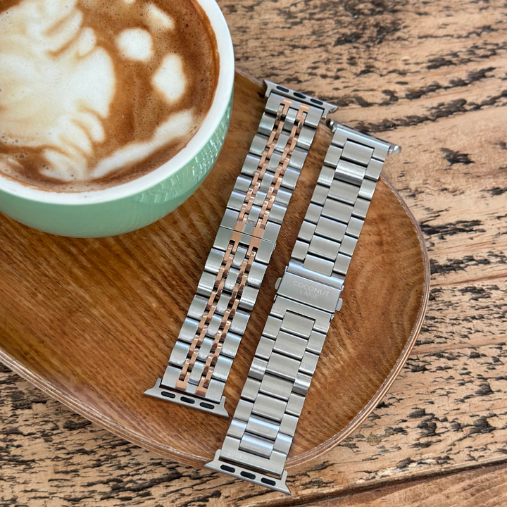 A green cup of latte with foam art sits on a wooden platter next to two interchangeable Coconut Lane Apple Watch straps. The straps are stainless steel; one has a mix of silver and rose gold links, and the other is the Stainless Steel Apple Watch Strap in Silver. The rustic wood surface adds to the cozy aesthetic.