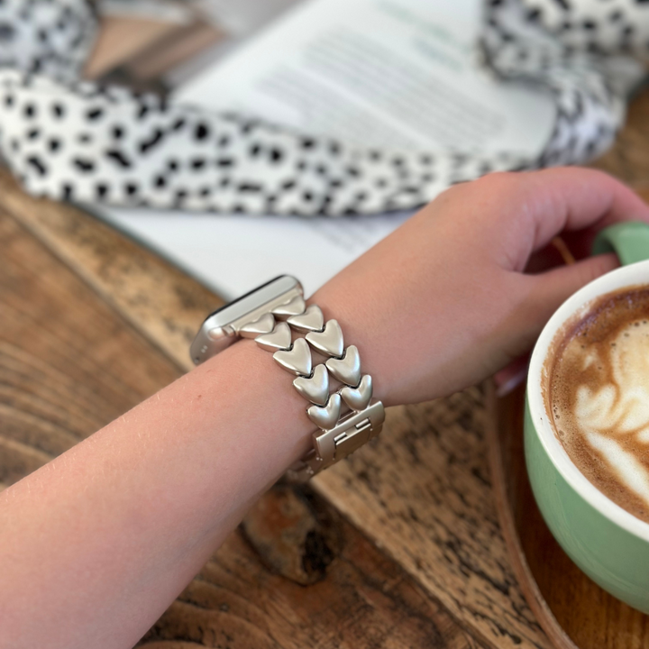 A person wearing a Hearts Chain Apple Watch Strap in Starlight by Coconut Lane on their wrist is holding a cup of coffee with latte art. A polka dot scarf and a magazine are visible in the background on a wooden table.