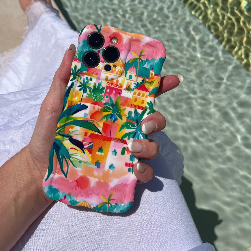 A person holding an iPhone with a colorful, tropical-themed Coconut Lane Wavy Phone Case - Portofino Dreams featuring palm trees and vibrant buildings. The person is wearing a white garment, and the background includes shimmering turquoise pool water and part of a white poolside lounger.