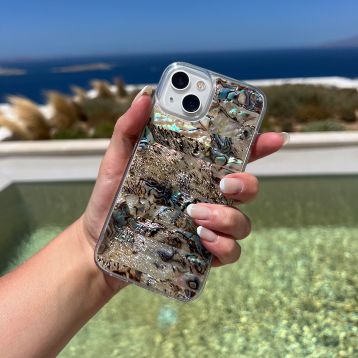 A hand with manicured nails holds a smartphone encased in the Coconut Lane Real Natural Shell Phone Case - Black, showcasing an iridescent abalone pattern. The background features a tranquil ocean view, a clear blue sky, and distant islands, evoking a serene coastal or beach setting.