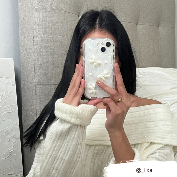 A person with long black hair wearing a white off-the-shoulder sweater holds a Coconut Lane Pearls & Bows Phone Case in front of their face, snapping a mirror selfie. A white textured headboard and white bed linen are visible in the background. A tag @_i.xa appears at the bottom right.