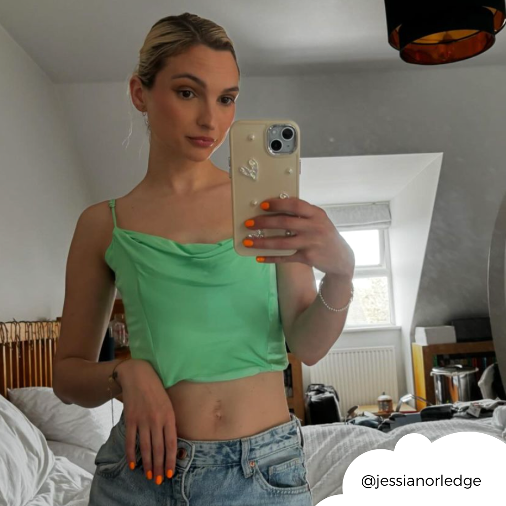 A person with blonde hair in a loose bun takes a mirror selfie in a bedroom with a slanted ceiling and a hanging black light fixture. They wear a green top and blue jeans, holding an extra protective Pearly Hearts Phone Case - Nude from Coconut Lane, adorned with a heart charm. The room is slightly cluttered with a bed and other items in the background.
