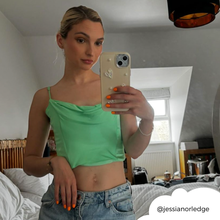 A person with blonde hair in a loose bun takes a mirror selfie in a bedroom with a slanted ceiling and a hanging black light fixture. They wear a green top and blue jeans, holding an extra protective Pearly Hearts Phone Case - Nude from Coconut Lane, adorned with a heart charm. The room is slightly cluttered with a bed and other items in the background.