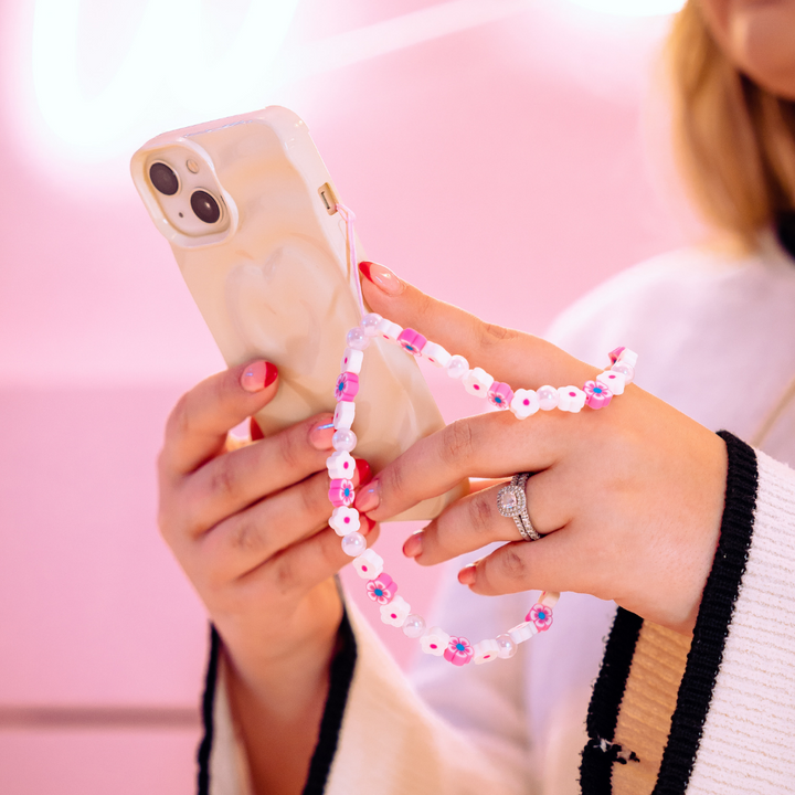 Close-up of a person holding an iPhone with Coconut Lane's Melting Heart Phone Case in Coconut Cream, adorned with a pink and white beaded strap. The individual, wearing a white sweater with black trim and a diamond ring on the ring finger of their left hand, is set against a slightly blurred pink background.