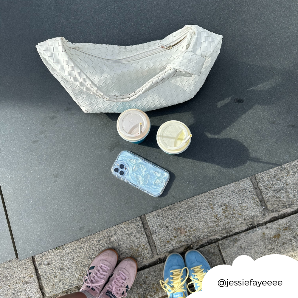 An overhead shot captures two pairs of feet—one in pink shoes, the other in blue and yellow shoes—a light-colored woven handbag, two takeaway drinks, and a smartphone encased in a Shimmer Blue Butterflies Phone Case by Coconut Lane on a stone surface. The Instagram handle @jessiefayeeee is written on the image.