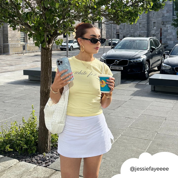 A woman is wearing a light yellow shirt and white skirt, holding a coffee cup in one hand and her phone with the Shimmer Blue Butterflies Phone Case from Coconut Lane in the other. She accessorizes with sunglasses, red earrings, and a white bag on her shoulder. She stands on a paved street with trees and cars in the background. Her Instagram handle "@jessiefayeeee" is visible in the corner.