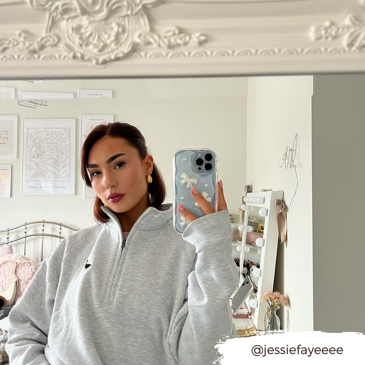 A woman stands in front of a mirror taking a selfie with her phone, adorned in the Pearls & Bows Phone Case by Coconut Lane. She is wearing a light grey zip-up sweatshirt and has long dark hair. Behind her, there is a decorated bedroom with framed pictures on the wall and a vanity with a mirror. Her Instagram handle, @jessiefayeeee, is written on the image.