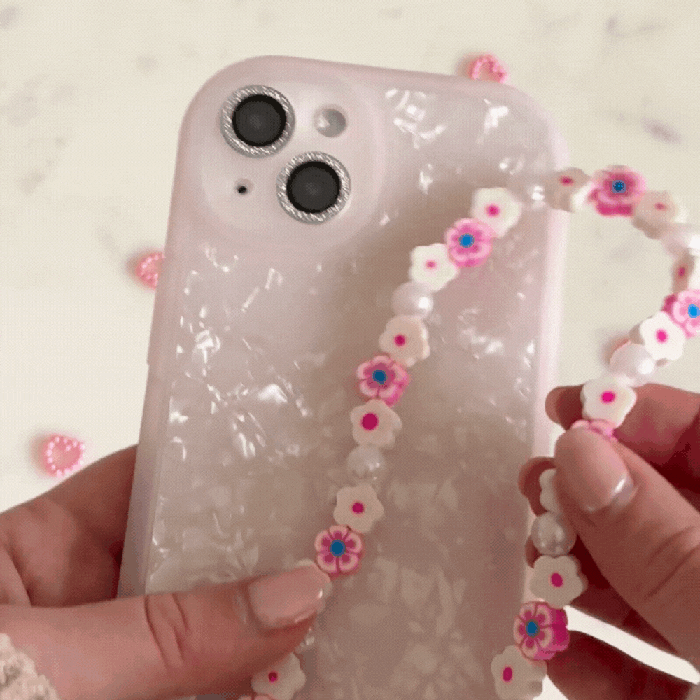 Someone is attaching the Coconut Lane Beaded Phone Strap - Flowers & Pearls, adorned with pink and white flower-shaped beads, to a textured white phone case. The phone features a dual-camera system, and the backdrop is a light surface sprinkled with decorative accessories.