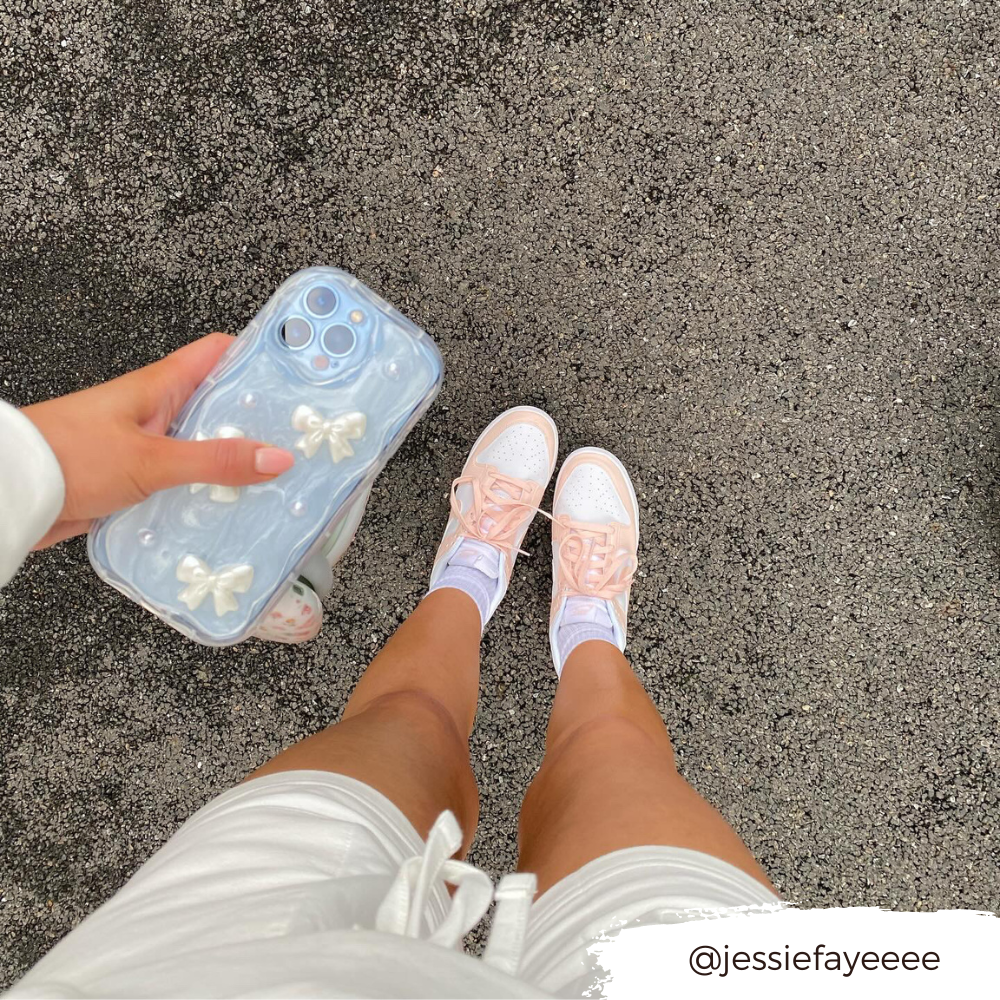 A person's legs are shown from above, wearing white shorts, white socks, and pastel pink sneakers. They are holding a phone with the Pearls & Bows Phone Case by Coconut Lane. The ground is an asphalt surface. The Instagram handle @jessiefayeeee is overlaid in the bottom right corner.