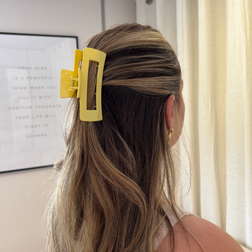 Woman with highlighted brown hair wears a yellow square Coconut Lane claw clip, standing indoors by a curtain.