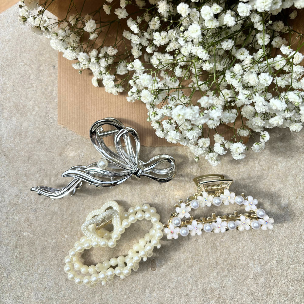 Three decorative hair clips with pearl adornments, including Coconut Lane's Pearl Heart Claw Clip, are displayed on a beige background next to a bouquet of small white flowers. These elegant hair accessories feature a silver bow design, the Pearl Heart Claw Clip from Coconut Lane, and a gold clasp with white floral decorations.