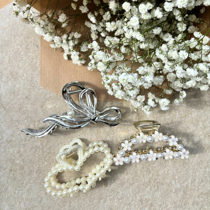 Three decorative hair clips with pearl adornments, including Coconut Lane's Pearl Heart Claw Clip, are displayed on a beige background next to a bouquet of small white flowers. These elegant hair accessories feature a silver bow design, the Pearl Heart Claw Clip from Coconut Lane, and a gold clasp with white floral decorations.