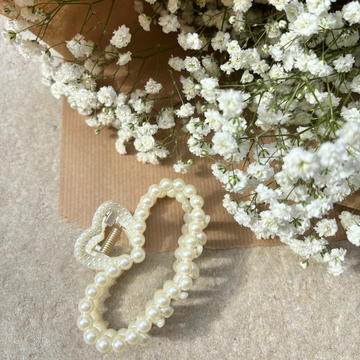 A Pearl Heart Claw Clip by Coconut Lane and a pearl hair tie are elegantly arranged next to a bouquet of delicate white baby's breath flowers on a textured beige surface, creating a soft and refined composition.