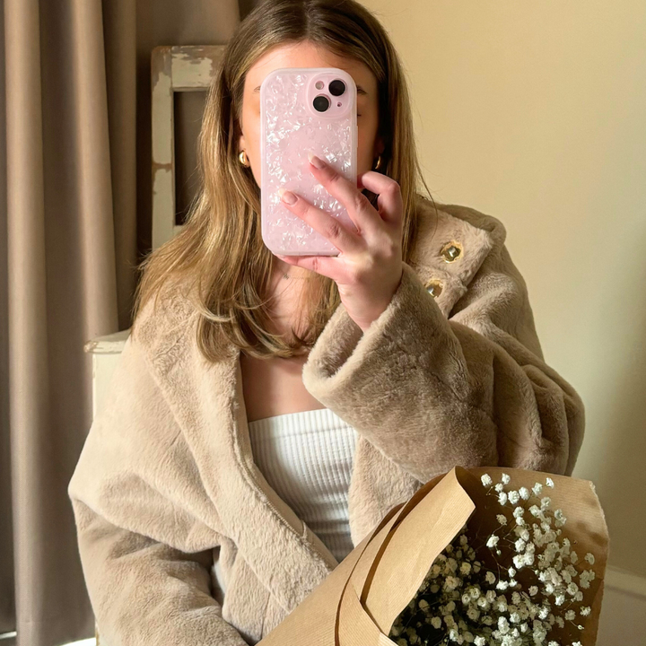 A person is taking a mirror selfie with an iPhone 15 that has a Coconut Lane Moonstone Pink Phone Case adorned with flowers. They are wearing a beige faux fur coat over a white top and holding a brown paper-wrapped bouquet of baby's breath flowers. The background shows a curtain and a piece of furniture.