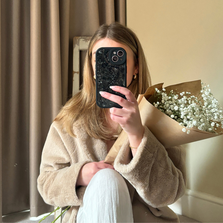 A person with long, light brown hair is sitting indoors and wearing a beige coat. They are holding a bouquet of white flowers wrapped in brown paper in one hand and covering their face with a Moonstone Black Phone Case from Coconut Lane in the other hand. A beige curtain and a wall are in the background.
