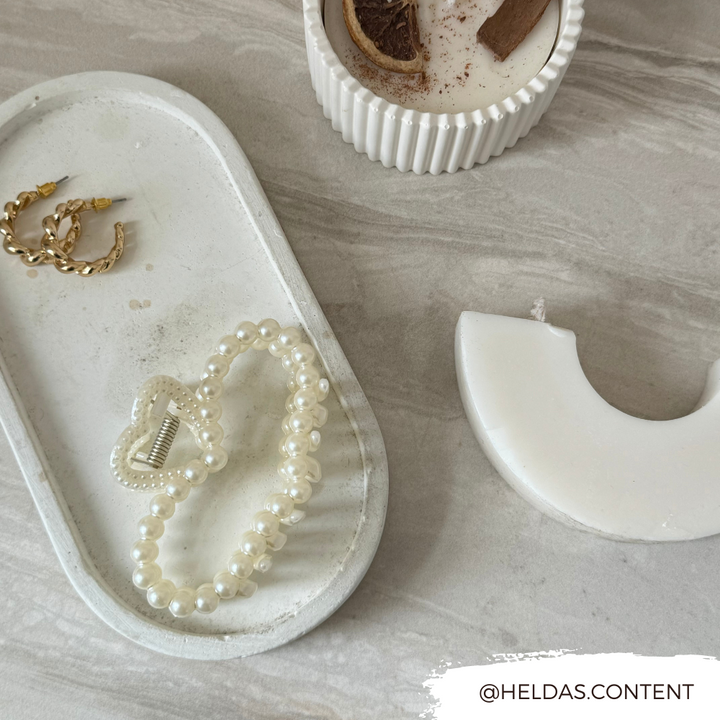 A flat lay photo on a marble surface features a pair of gold hoop earrings, a bracelet with pearl beads and a heart-shaped clasp on a white ceramic tray, a candle in a ribbed container, and an elegant Pearl Heart Claw Clip from Coconut Lane. Text at the bottom right reads "@HELDAS.CONTENT".