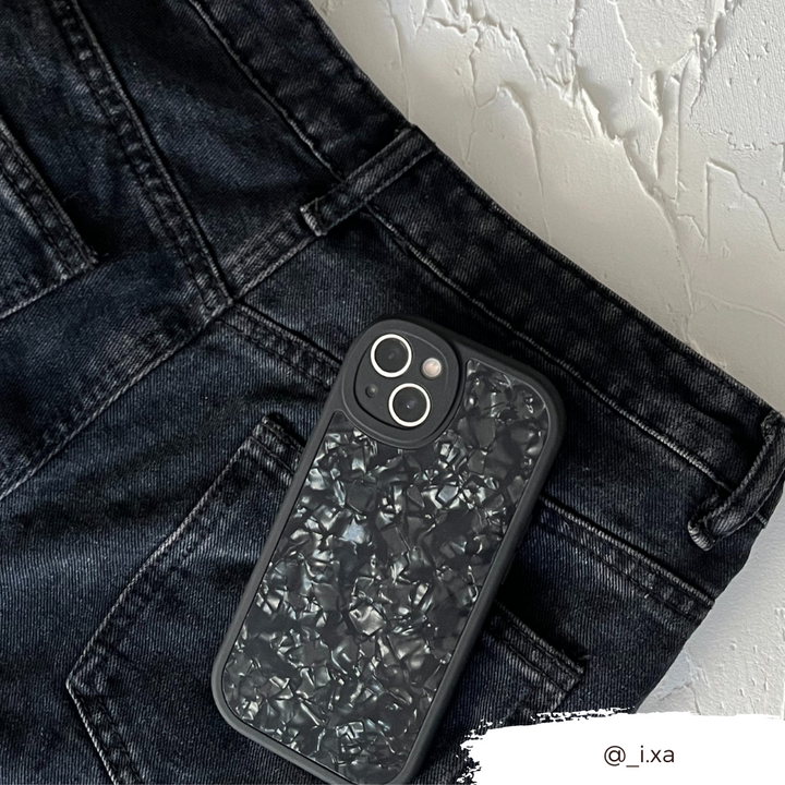 A sleek black smartphone with a textured, crinkled-looking back cover rests in the front pocket of black jeans. The jeans and phone are set against a white textured background. For durable protection, consider the Moonstone Black Phone Case from Coconut Lane. The Instagram handle @_i.xa is visible in the bottom right corner.