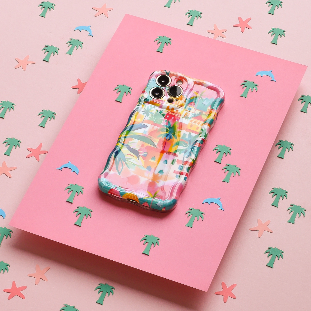 The Wavy Phone Case - Portofino Dreams by Coconut Lane, with its Italian seaside theme and colorful pastel patterns, is placed on a pink surface adorned with small illustrations of palm trees, starfish, and dolphins. This extra protective case adds a lively touch to the overall beachy design.