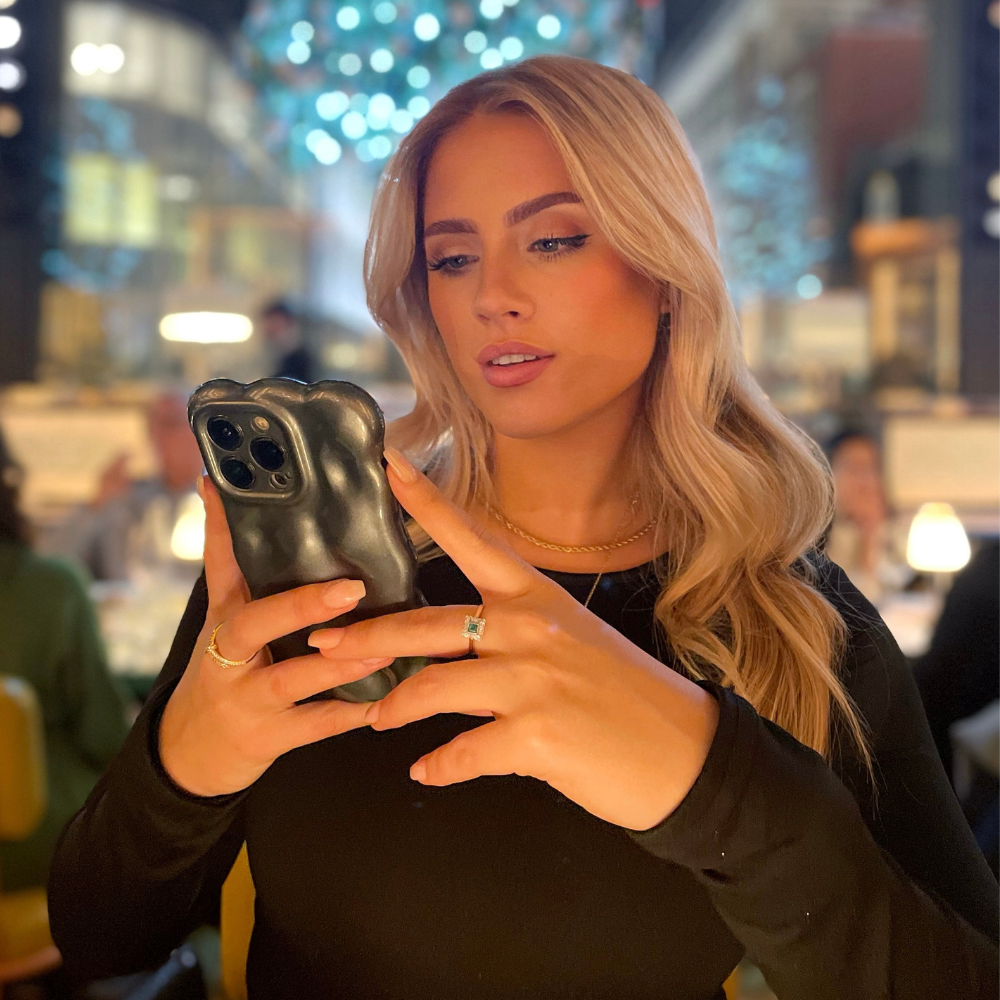 A woman with long blonde hair, wearing a black top and gold necklace, is looking at her phone in an elegantly lit restaurant where the lights are softly blurred and people are visible in the background. She appears focused as her fingers interact with the Coconut Lane 3D Pearl Phone Case - Grey on her iPhone.