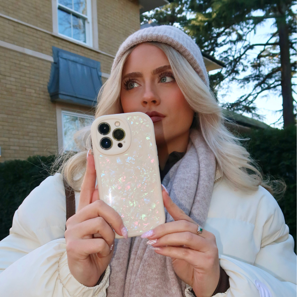 A person with long blonde hair, wearing a beige beanie, scarf, and white jacket, holds a Moonstone Pearl Phone Case by Coconut Lane in front of their face. They are standing outdoors in front of a house with green shrubs and trees in the background.