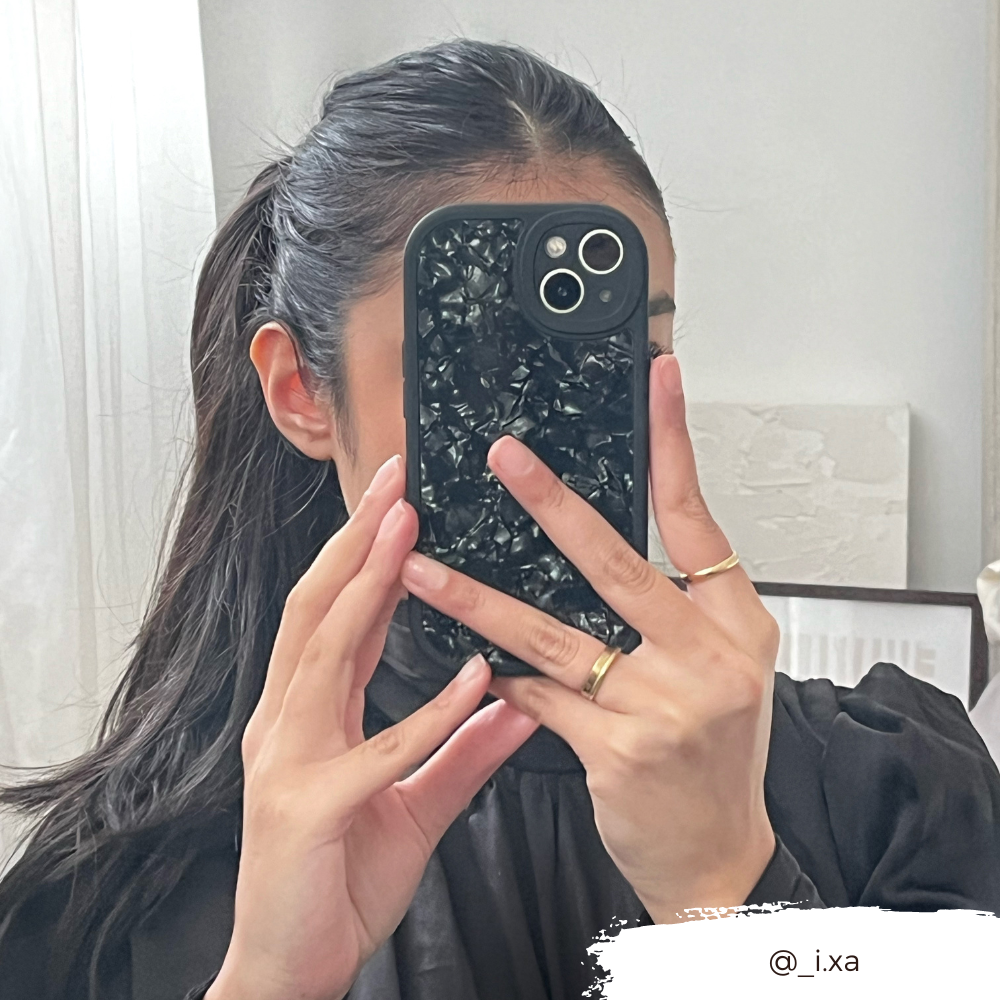 A person with dark hair in a ponytail takes a mirror selfie. They are holding an iPhone15 with the Moonstone Black Phone Case from Coconut Lane, which offers durable protection and partially covers their face. They wear a black top and several rings on their fingers. The Instagram handle @_i.xa is visible in the bottom right corner.