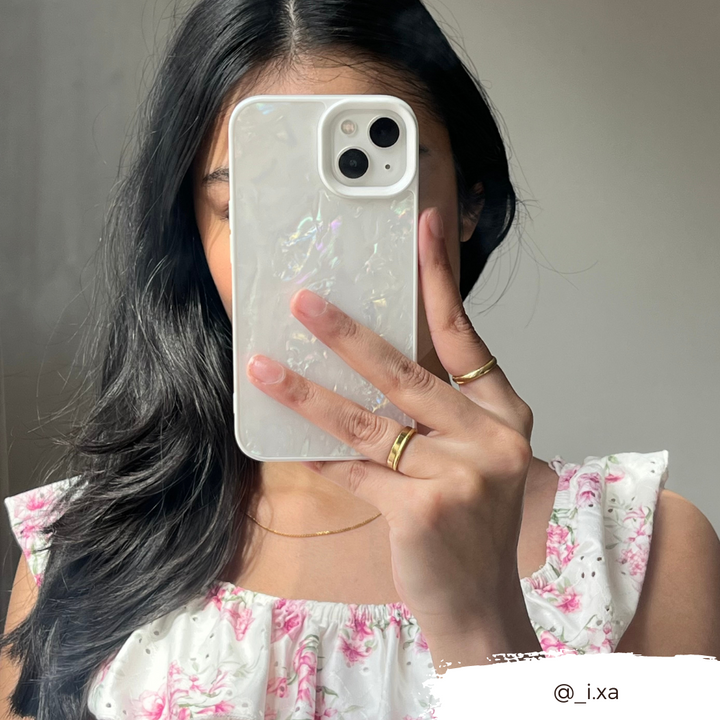 A woman with long black hair holds a white phone with the Coconut Lane Mother of Pearl Phone Case in front of her face, obscuring it. She is wearing a white dress adorned with pink floral patterns and multiple gold rings. The background is neutral, and the handle @_i.xa appears in the bottom right corner.